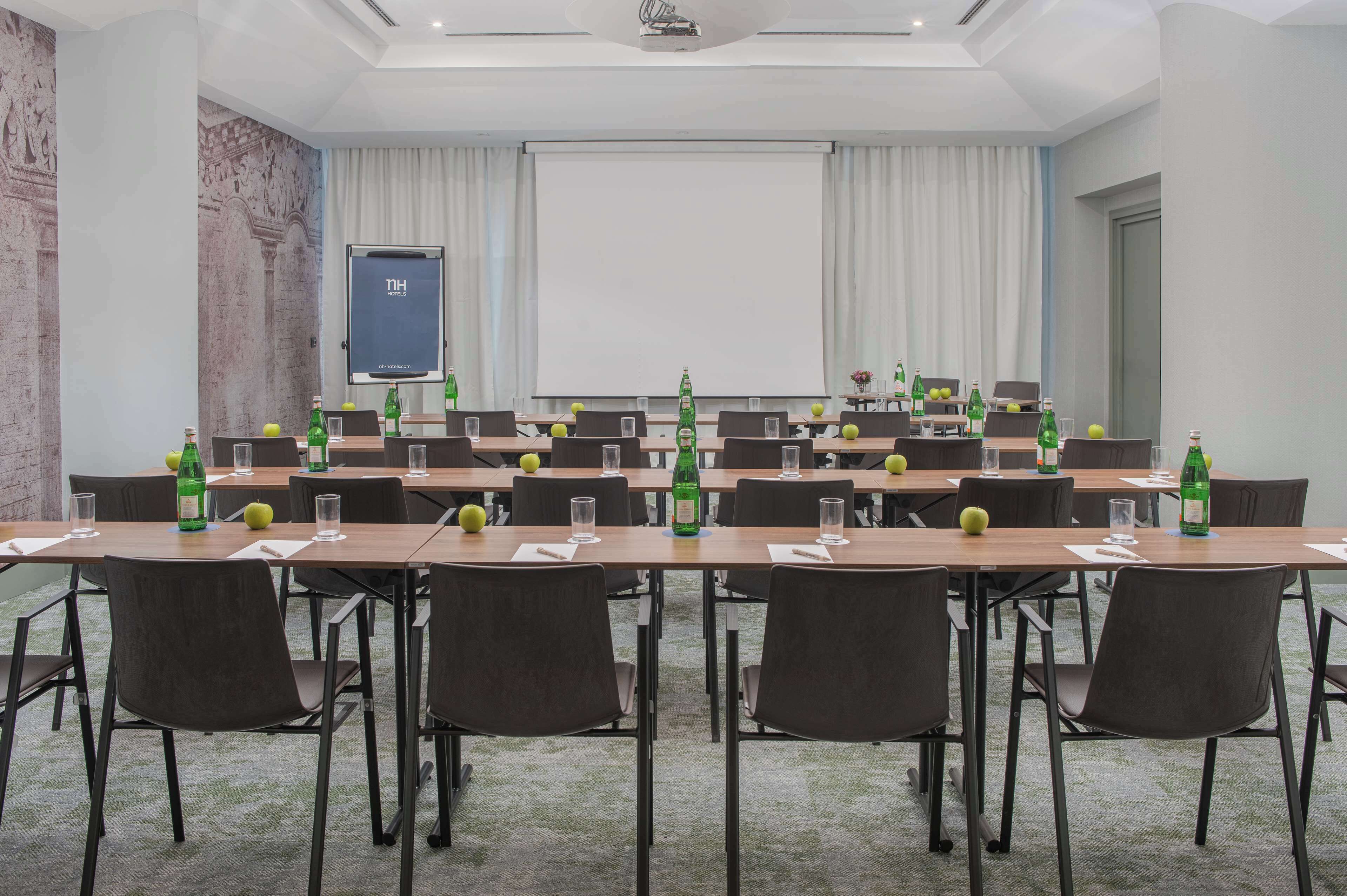 Una sala conferenze con un lungo tavolo, sedie, uno schermo per proiettore e bottiglie verdi sul tavolo.