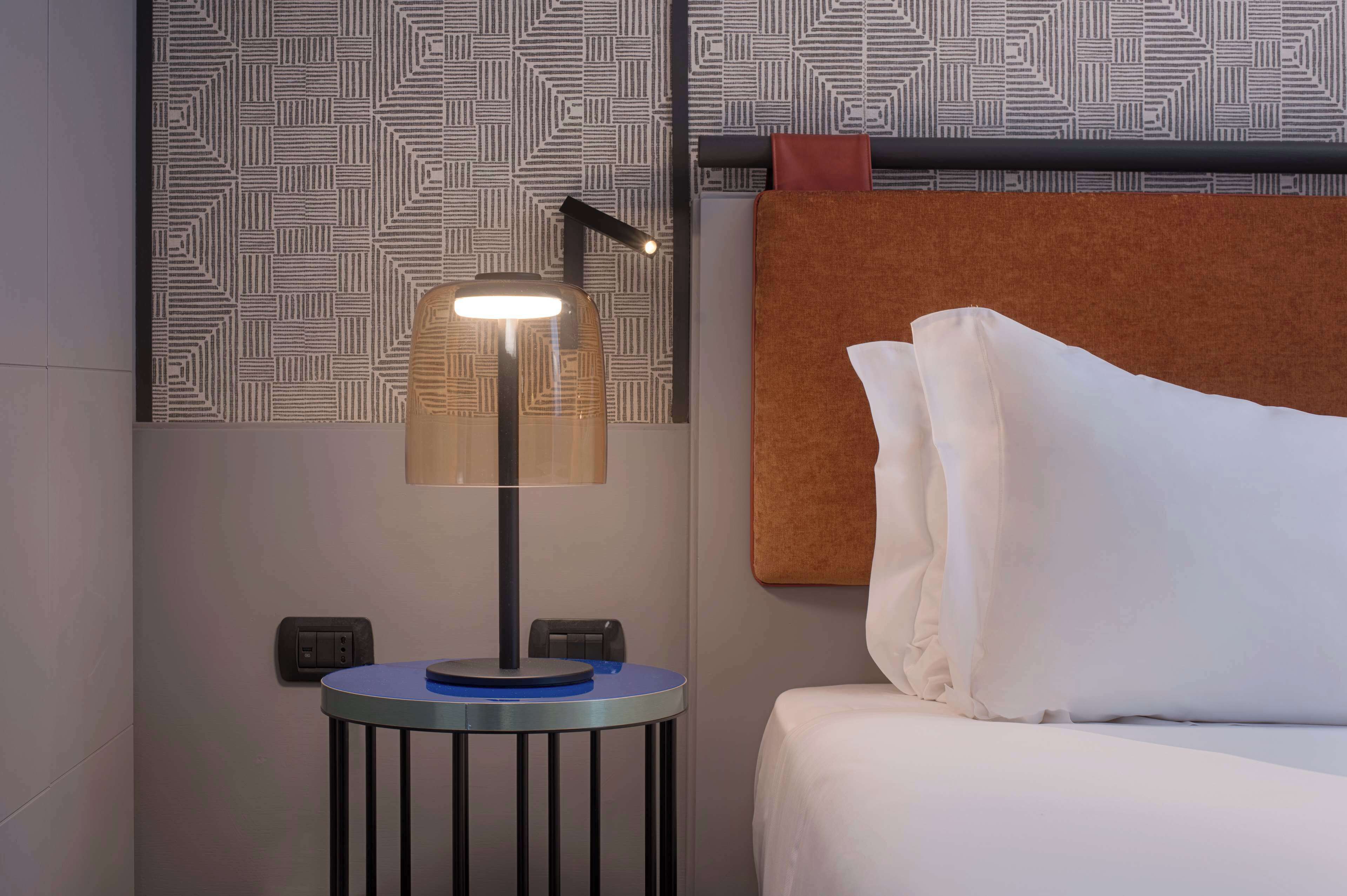 Ein ordentlich bezogenes Bett mit weißen Leinen, ein kleiner runder Tisch mit einer Lampe und eine Wand mit Muster im Hintergrund.