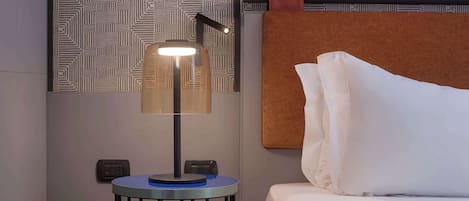 Ein ordentlich bezogenes Bett mit weißen Leinen, ein kleiner runder Tisch mit einer Lampe und eine Wand mit Muster im Hintergrund.