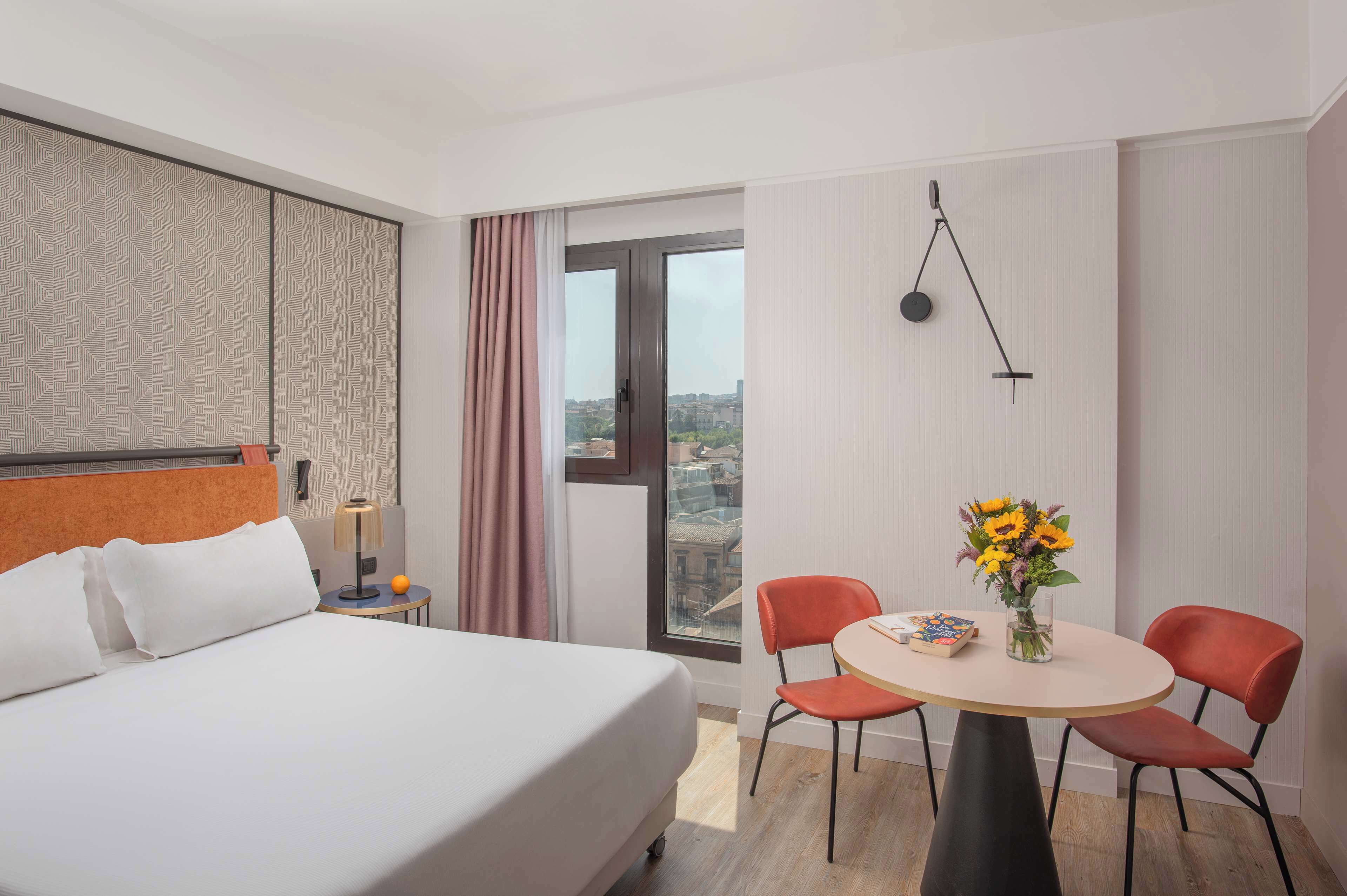 Una habitación de hotel con una cama, una mesa de comedor con sillas, un jarrón con flores y vistas a través de una ventana.