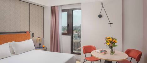 Una habitación de hotel con una cama, una mesa de comedor con sillas, un jarrón con flores y vistas a través de una ventana.