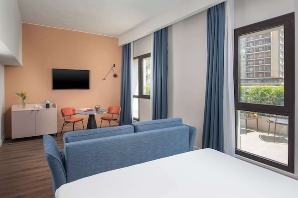 A modern hotel room with a large bed, a flat-screen TV mounted on the wall, a small table with chairs, and a view of the city through large windows. - NH Catania Centro (Catania)