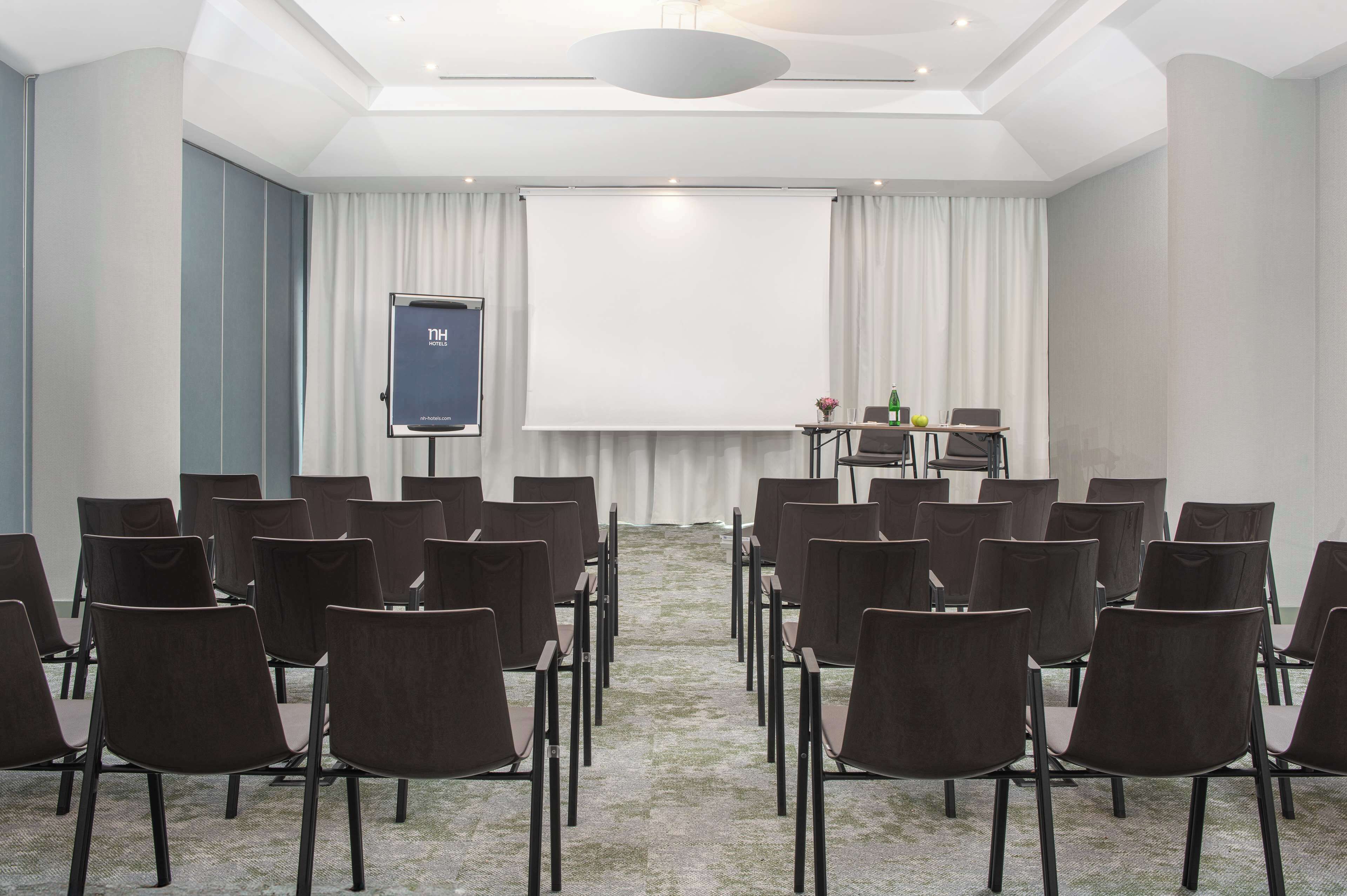 Une salle de conférence avec des rangées de chaises disposées face à un écran de projection, une table sur laquelle se trouvent un ordinateur portable et une bouteille, et une grande fenêtre avec des rideaux.