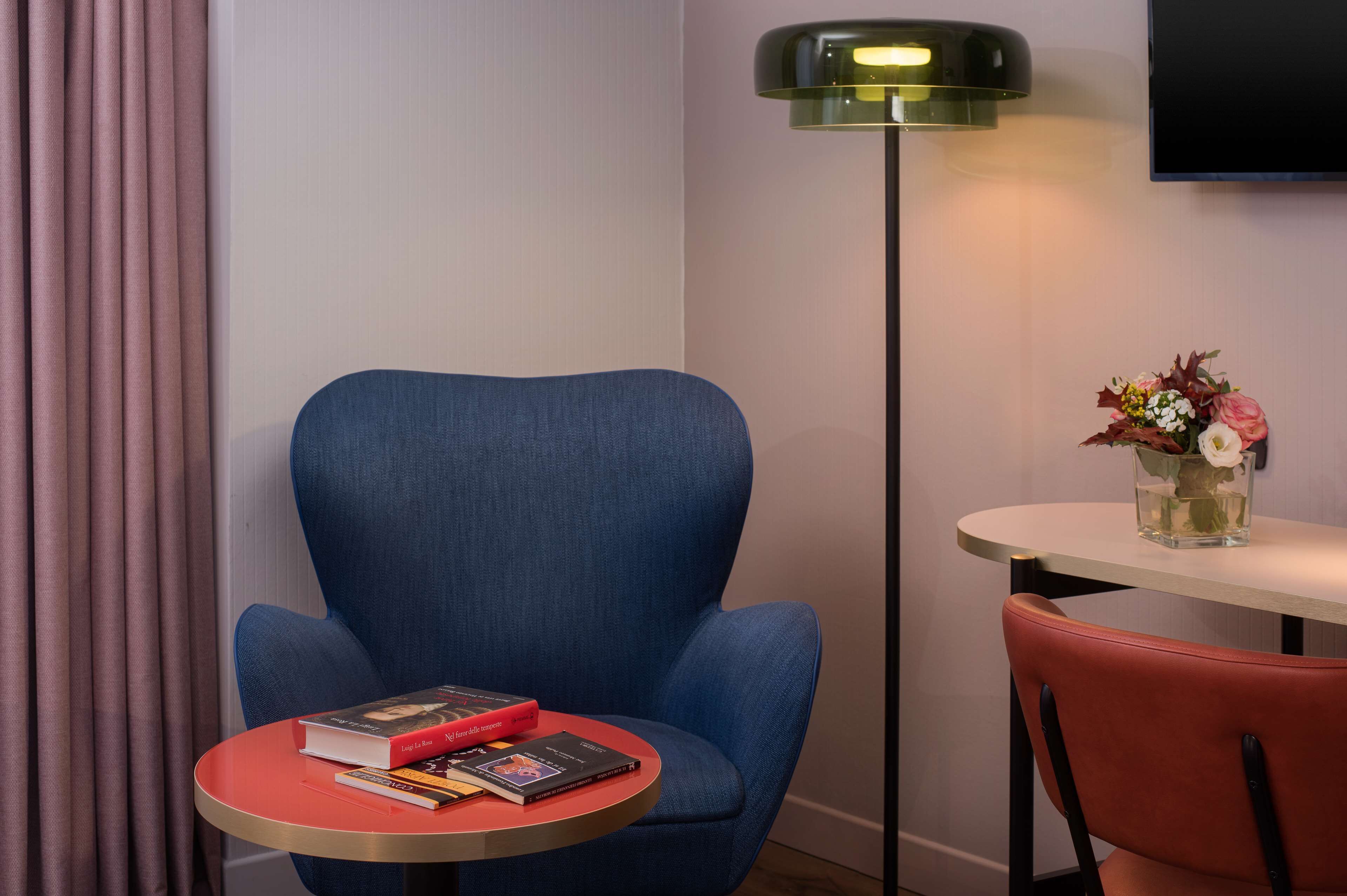 Una stanza con una poltrona blu, un tavolino rotondo con dei libri, una sedia rossa, una lampada da terra e un vaso con fiori.