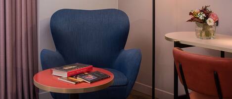 Una stanza con una poltrona blu, un tavolino rotondo con dei libri, una sedia rossa, una lampada da terra e un vaso con fiori.