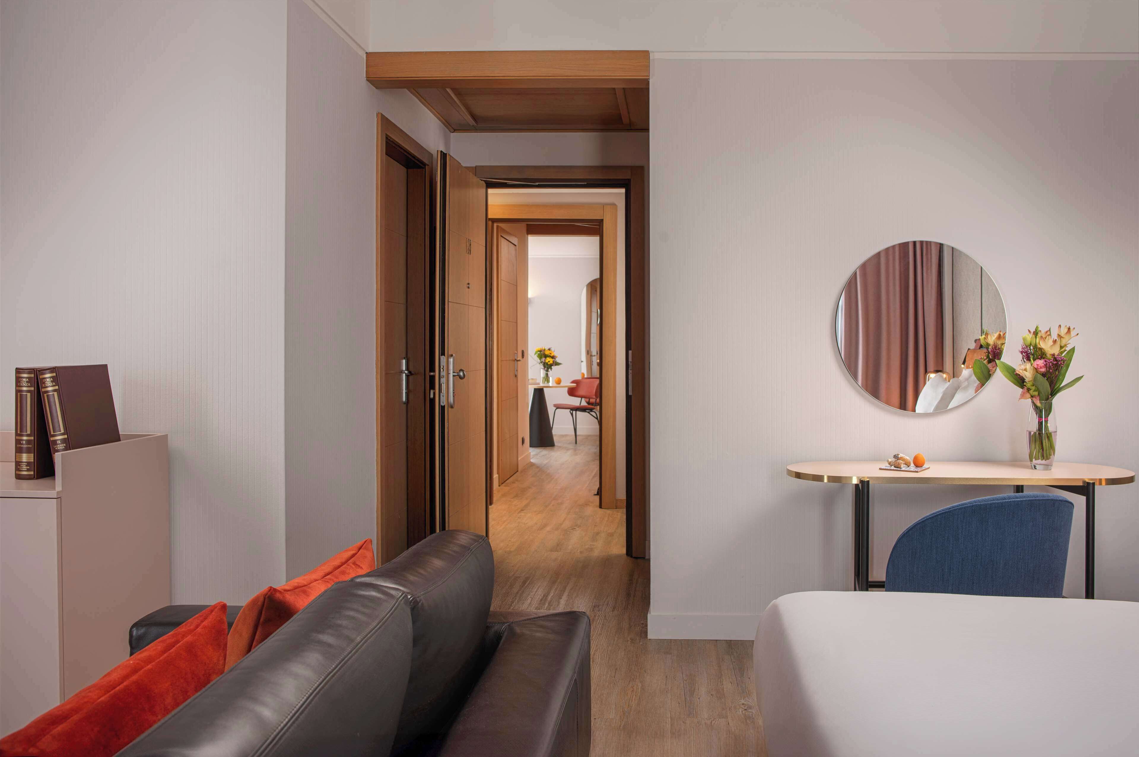 A hotel room with a bed, a desk with a mirror, a sofa, and a view of a hallway leading to a dining area.