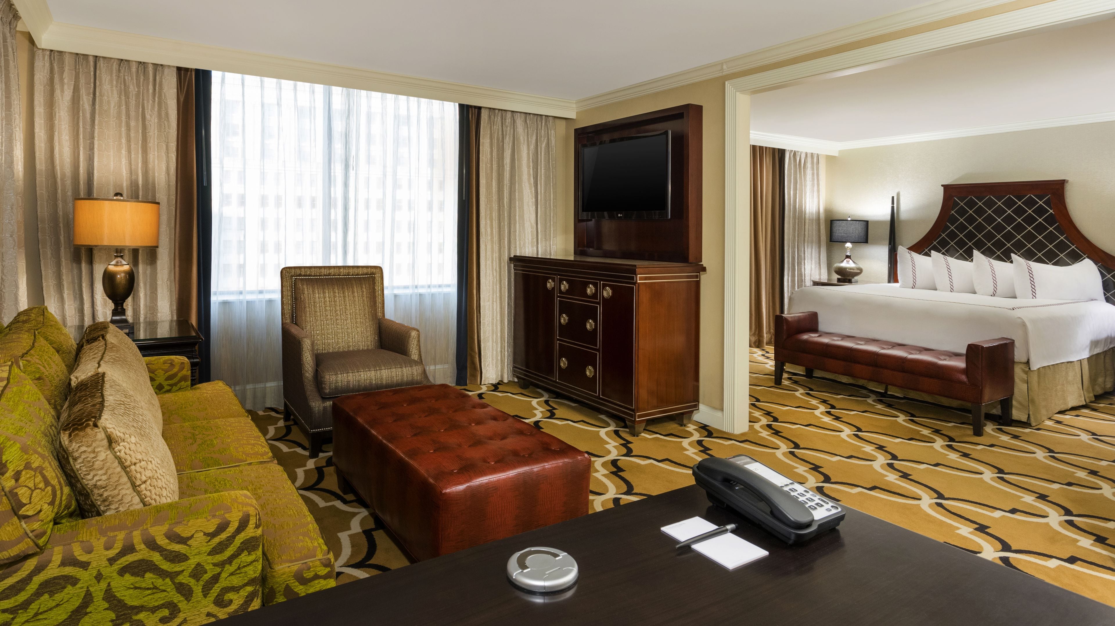 A hotel room with a sofa, armchair, coffee table, desk with a telephone, and a bed with a headboard.