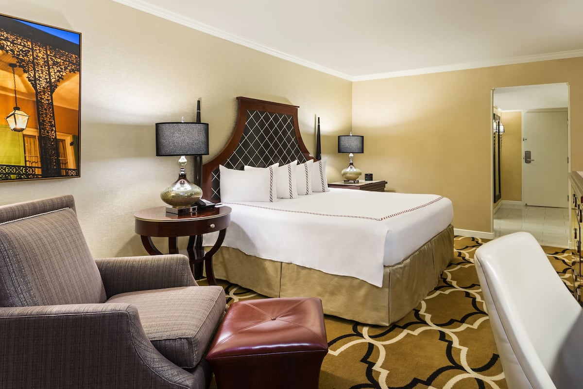 A hotel room with a bed, a desk, a chair, a lamp, and a painting on the wall.