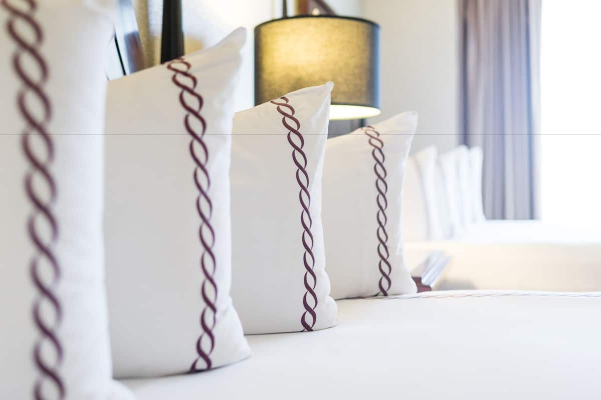 A hotel room with neatly arranged white pillows with a chain pattern, a lamp, and a window with curtains.