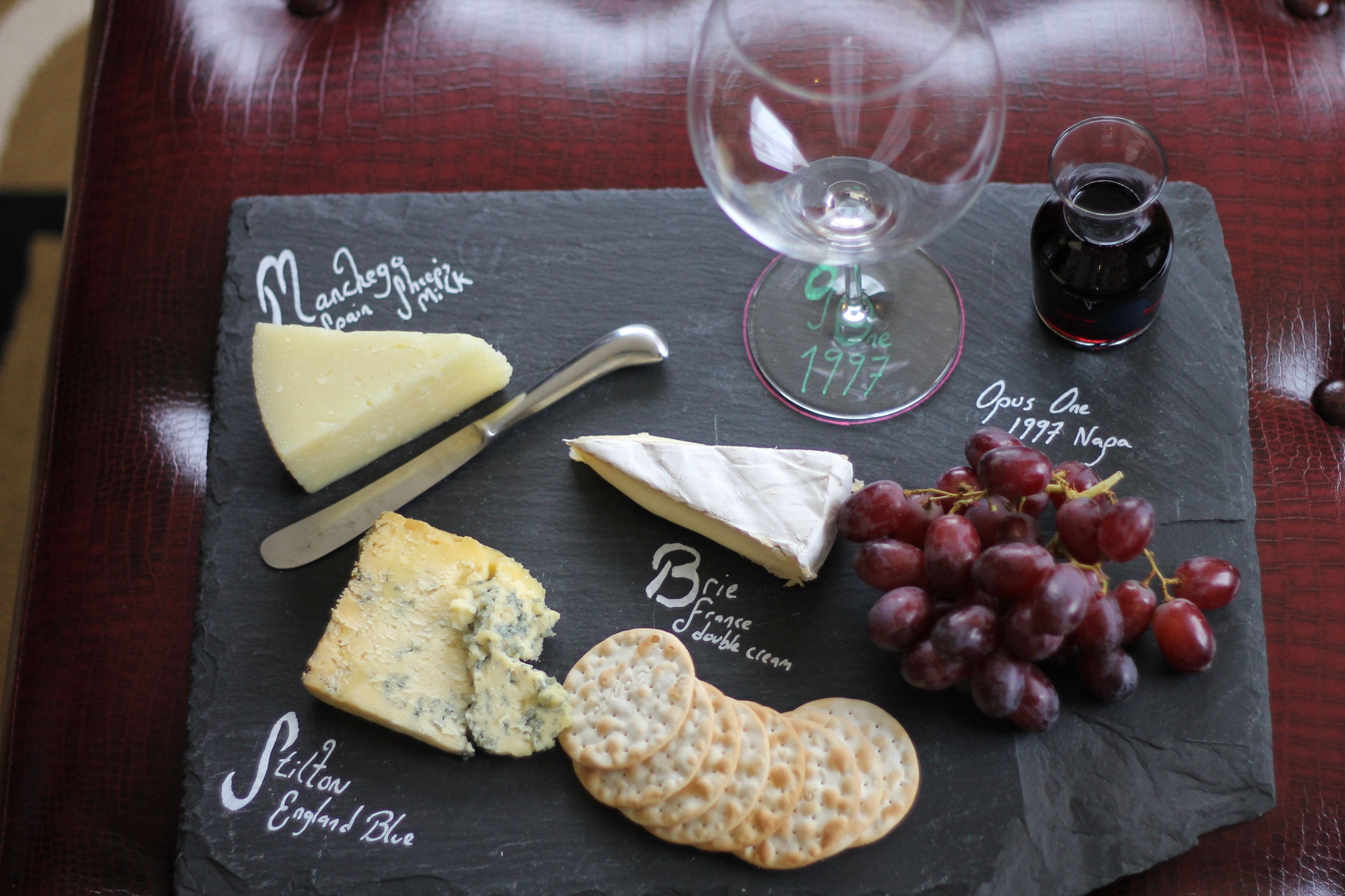 A slate board with various cheeses, crackers, and grapes, accompanied by a wine glass and a bottle of wine.
