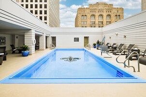 A rooftop pool with a fleur-de-lis logo, surrounded by lounge chairs and potted plants, with a view of a historic building in the background.