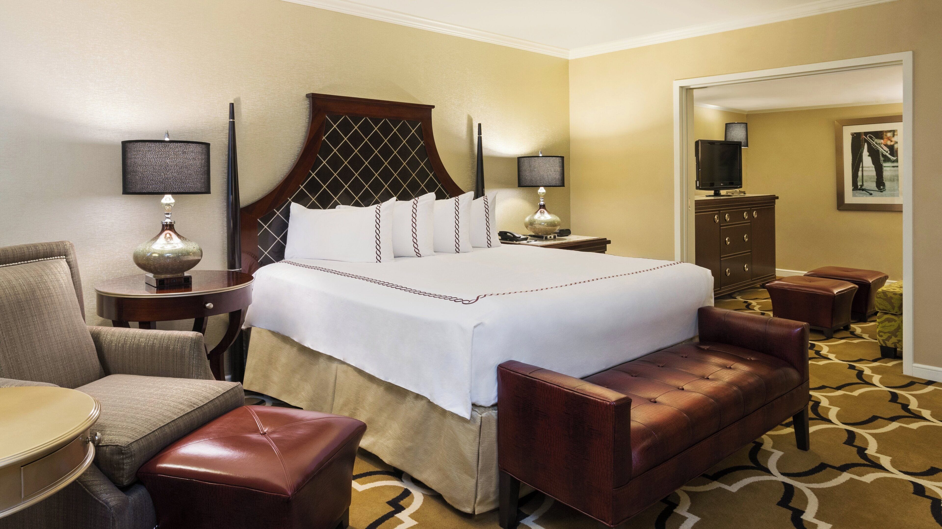 A hotel room with a large bed, a sitting area with a chair and ottoman, a desk, and a dresser.