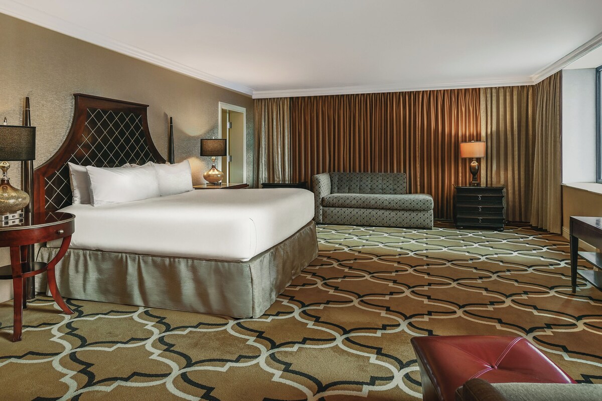 A hotel room with a large bed, a sofa, a TV, and a patterned carpet.