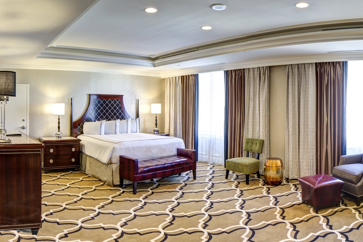 A hotel room with a large bed, a sitting area with a sofa and chairs, a desk with a lamp, and a large window with curtains.