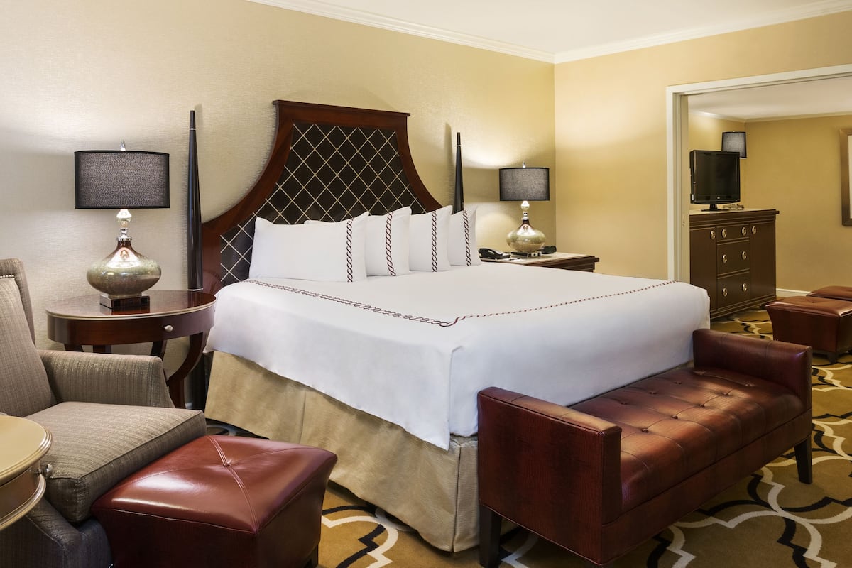 A hotel room with a large bed, a sitting area with a chair and ottoman, a desk with a lamp, a dresser, and a television.