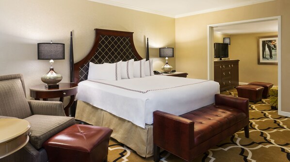 Una habitación de hotel con una cama grande, una zona de estar con un sillón y reposapiés, un escritorio con lámpara, un aparador y un televisor.