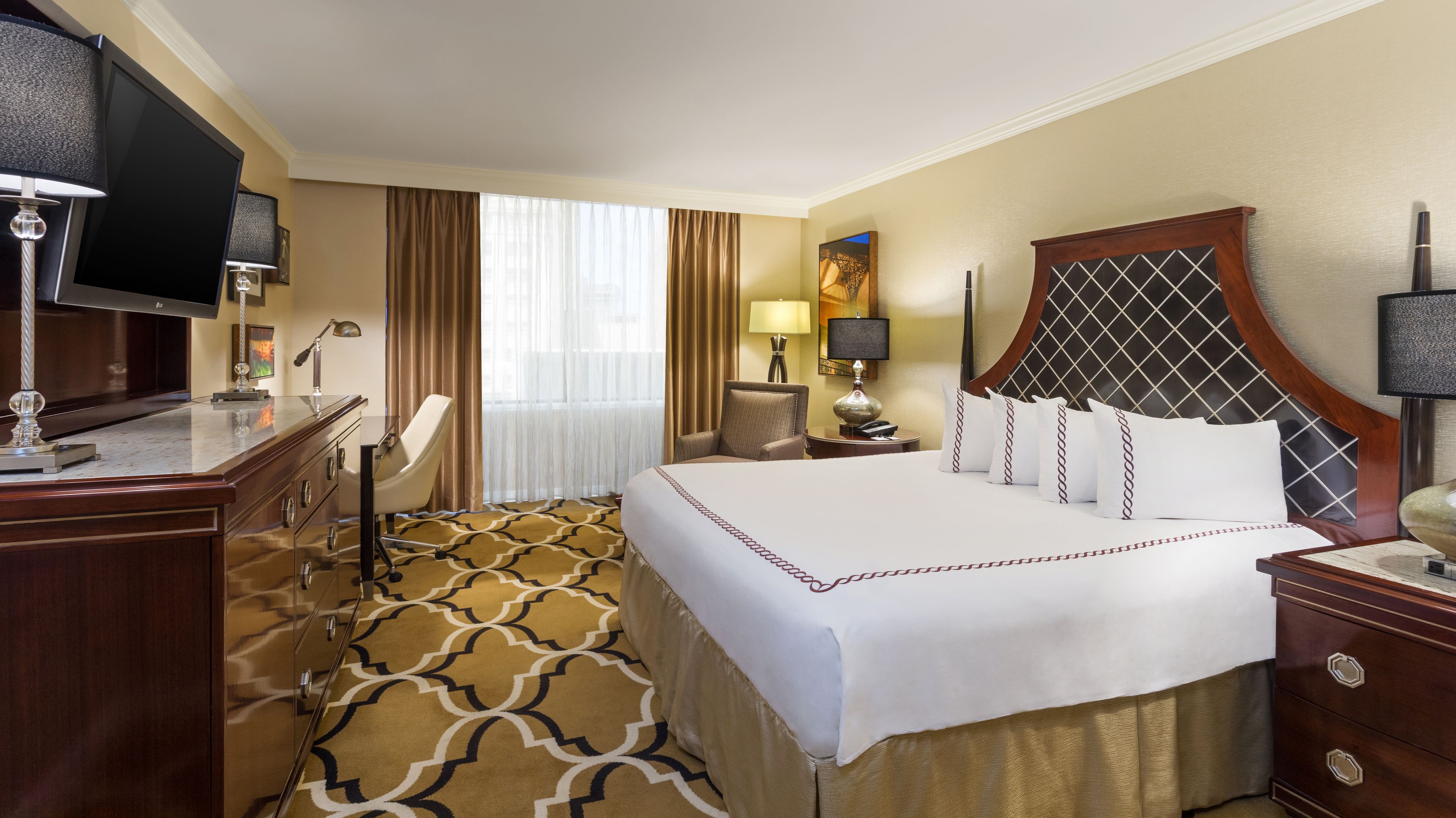 A hotel room with a large bed, a desk with a flat-screen TV, a chair, a nightstand with a lamp, and a window with curtains.