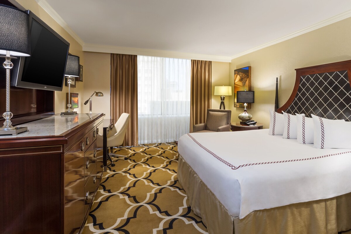 A hotel room with a large bed, a desk with a flat-screen TV, a chair, a nightstand with a lamp, and a window with curtains.