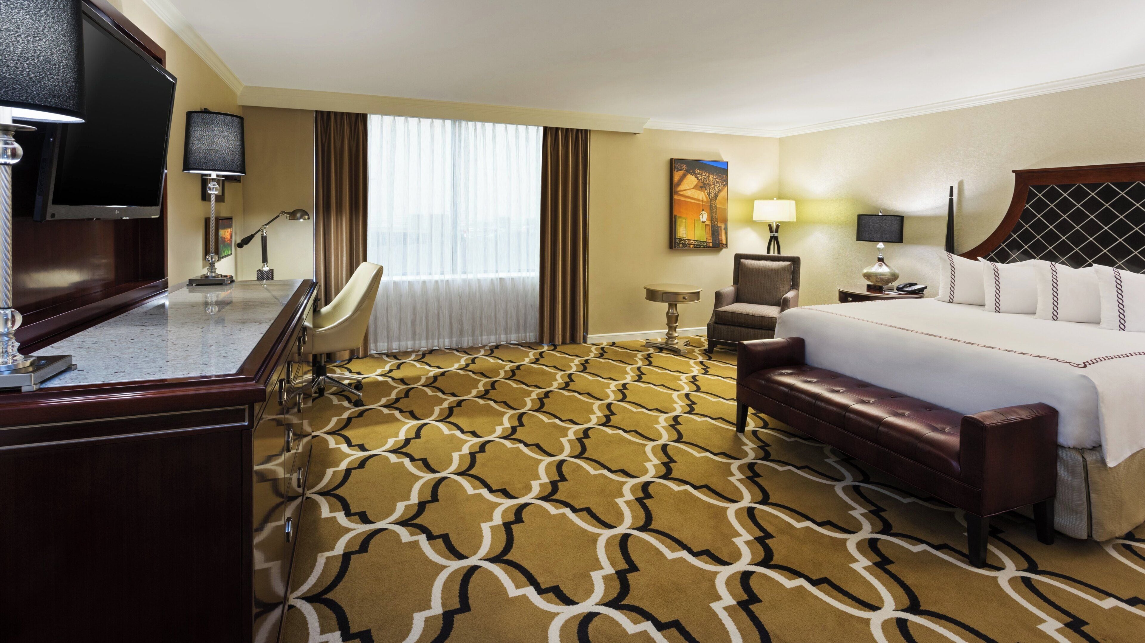 Une chambre d’hôtel avec un grand lit, un bureau doté d’un téléviseur à écran plat, une chaise, une petite table et un tapis à motifs géométriques.