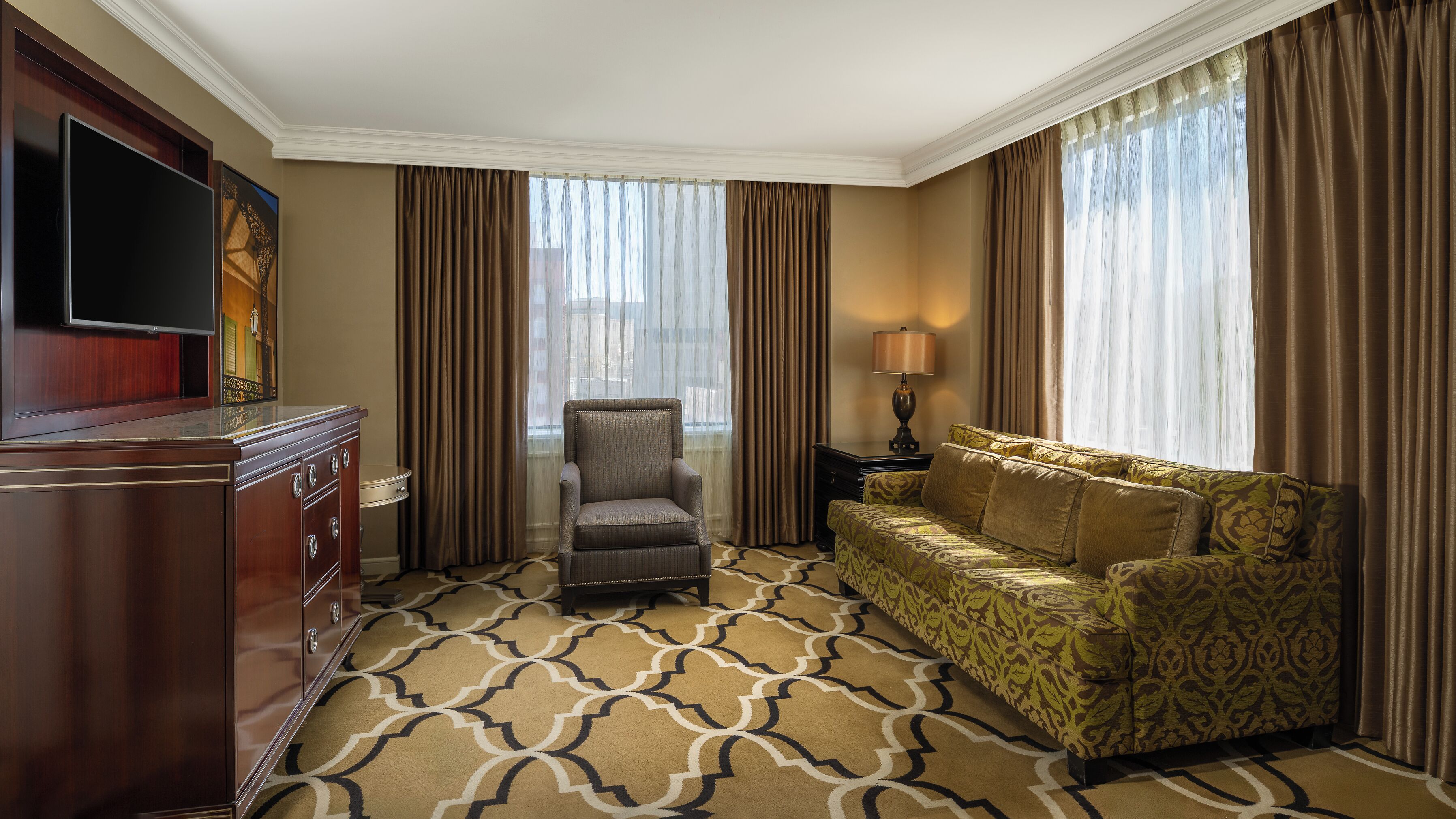 A hotel room with a flat-screen TV, a sofa, a chair, a lamp, and a patterned carpet.