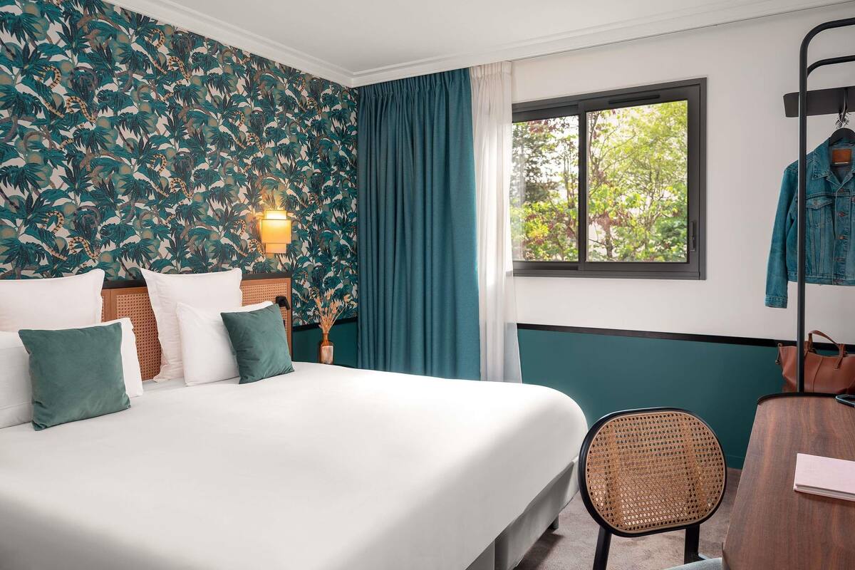 A hotel room with a bed, a desk, a chair, a window with a view of trees, and a wall with a leafy pattern.
