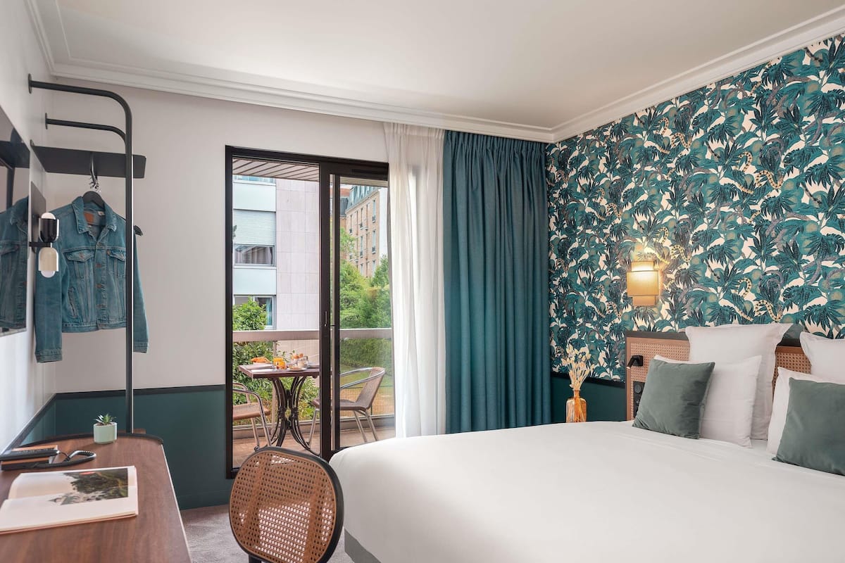 A hotel room with a bed, a desk with a chair, a hanging rack with clothes, and a view of the outside.