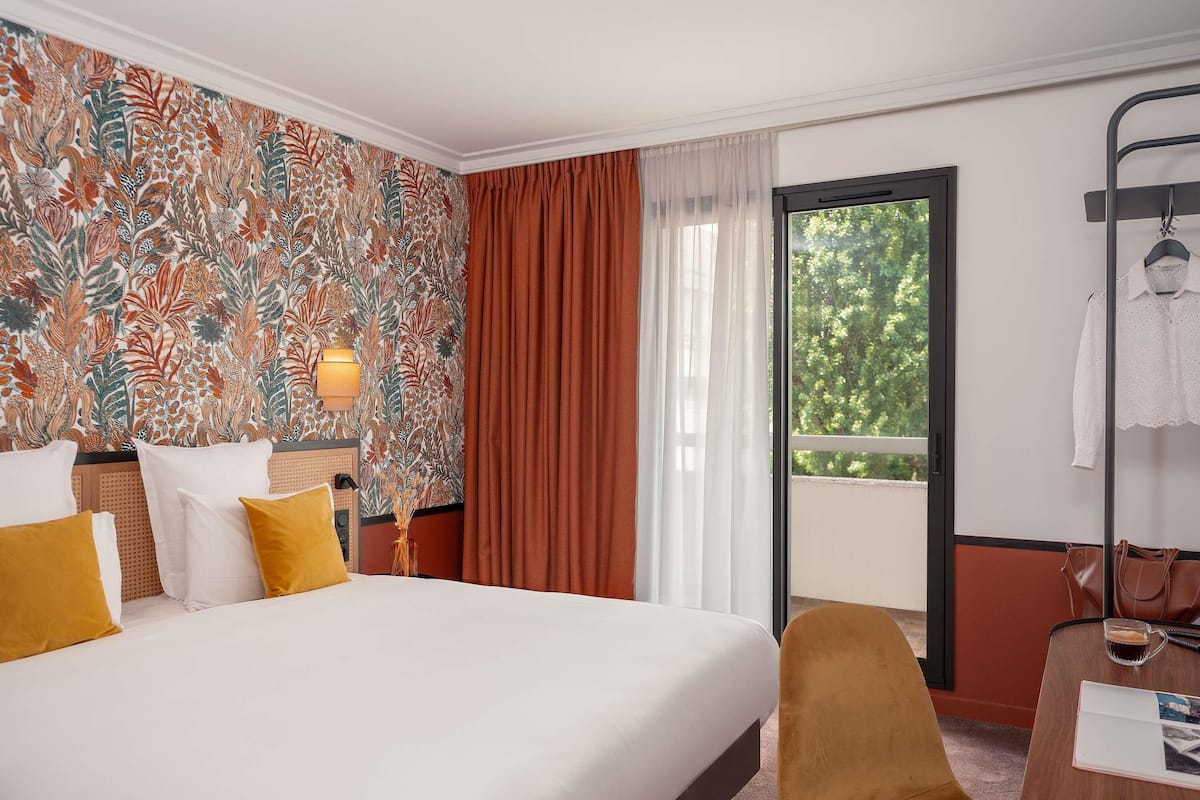 A hotel room with a large bed, a desk with a chair, a window with a view of trees, and a wall with a floral pattern.