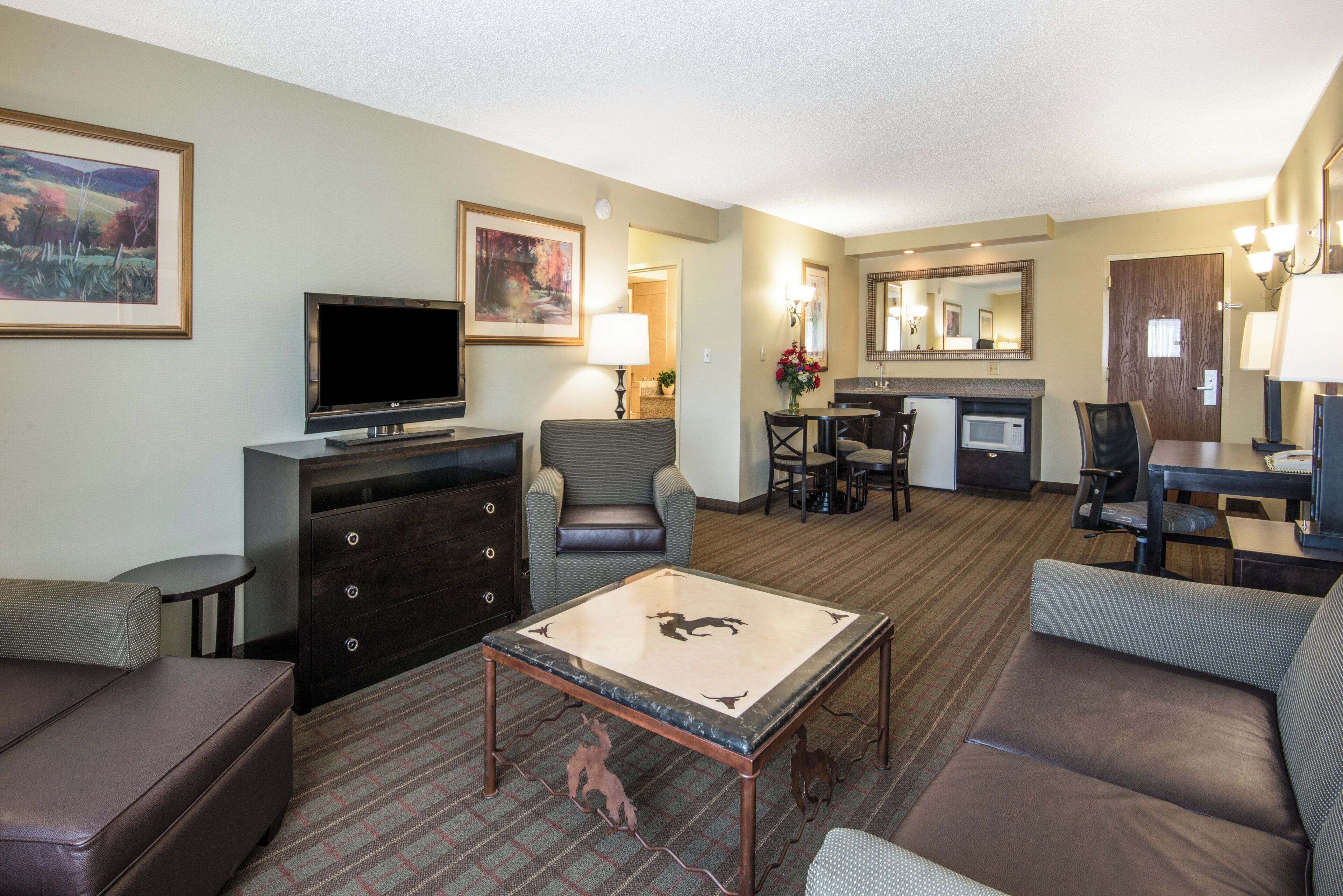 Une chambre d’hôtel équipée d’un téléviseur à écran plat, d’un canapé, d’une table basse, d’un coin repas avec une table et des chaises, d’un coin cuisine avec un four à micro-ondes, et d’une salle de bain avec un miroir et un lavabo.