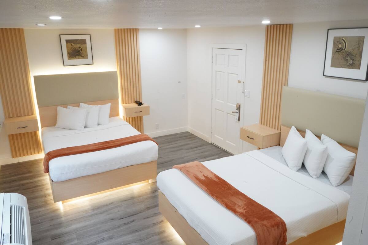 A hotel room with two beds, a wooden floor, a white door, and a framed picture on the wall.