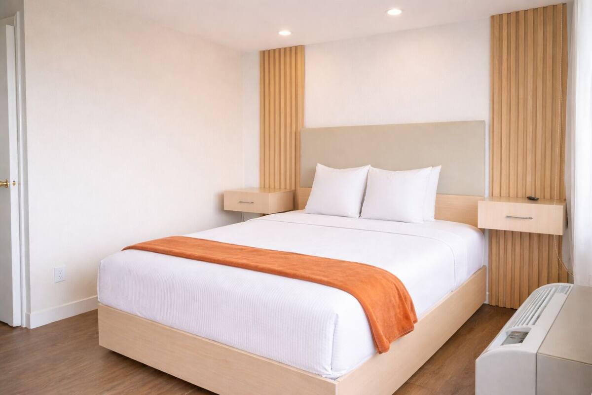A modern hotel room with a large bed, wooden headboard, bedside tables, and a built-in heater.