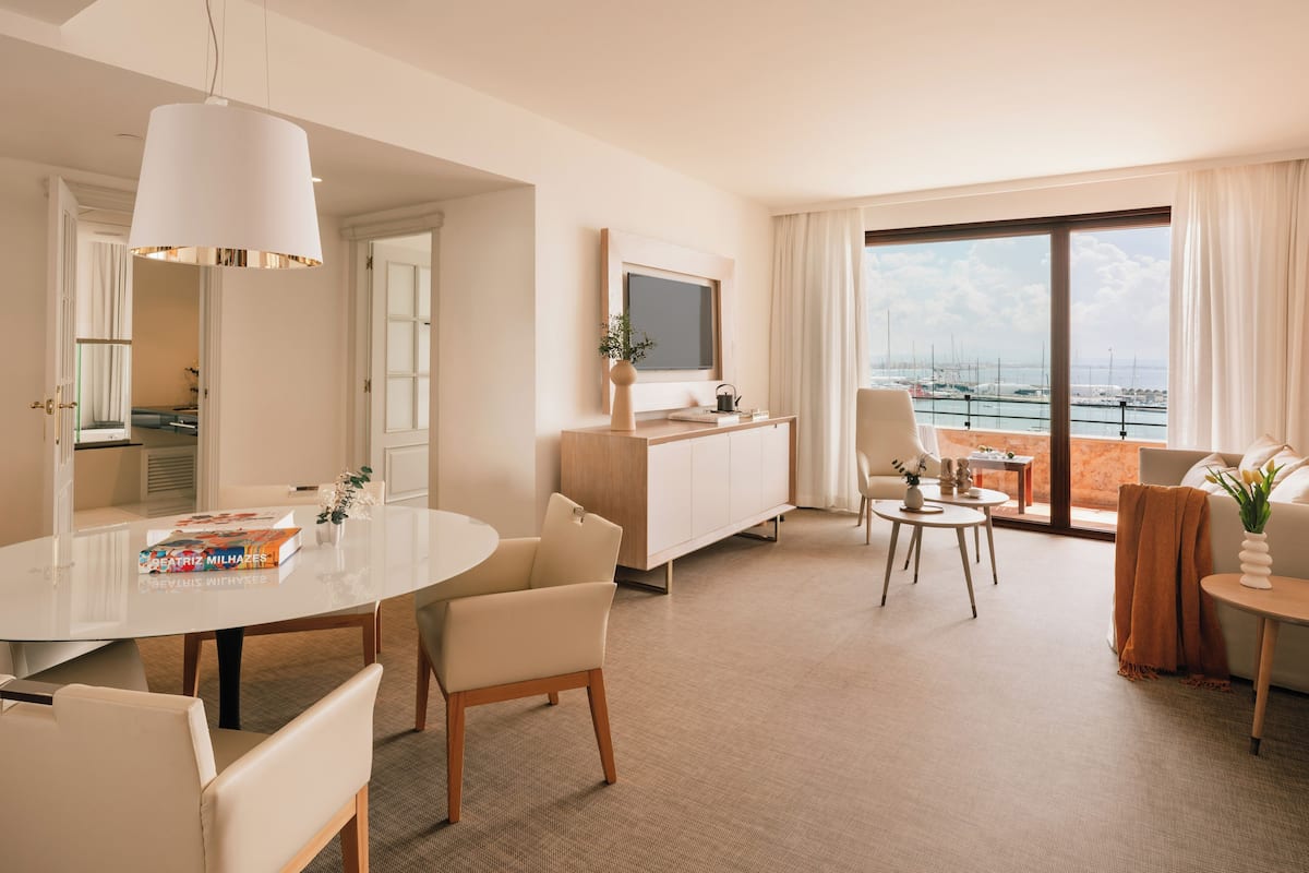 Una habitación de hotel moderna con zona de comedor, sala de estar y una gran ventana con vista al puerto deportivo.