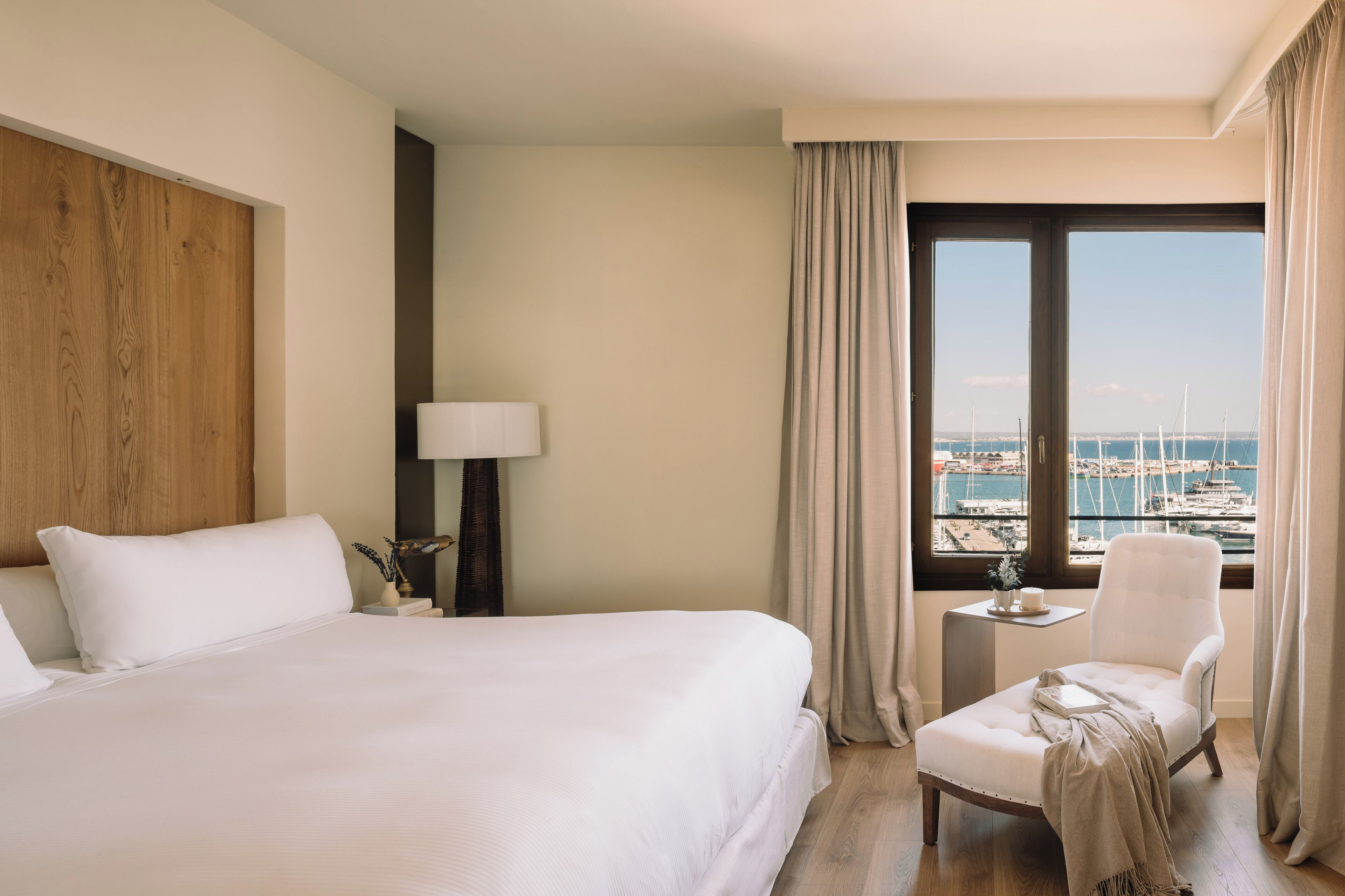 Habitación de hotel con una cama grande, una silla, una mesita con lámpara y vistas al puerto deportivo.