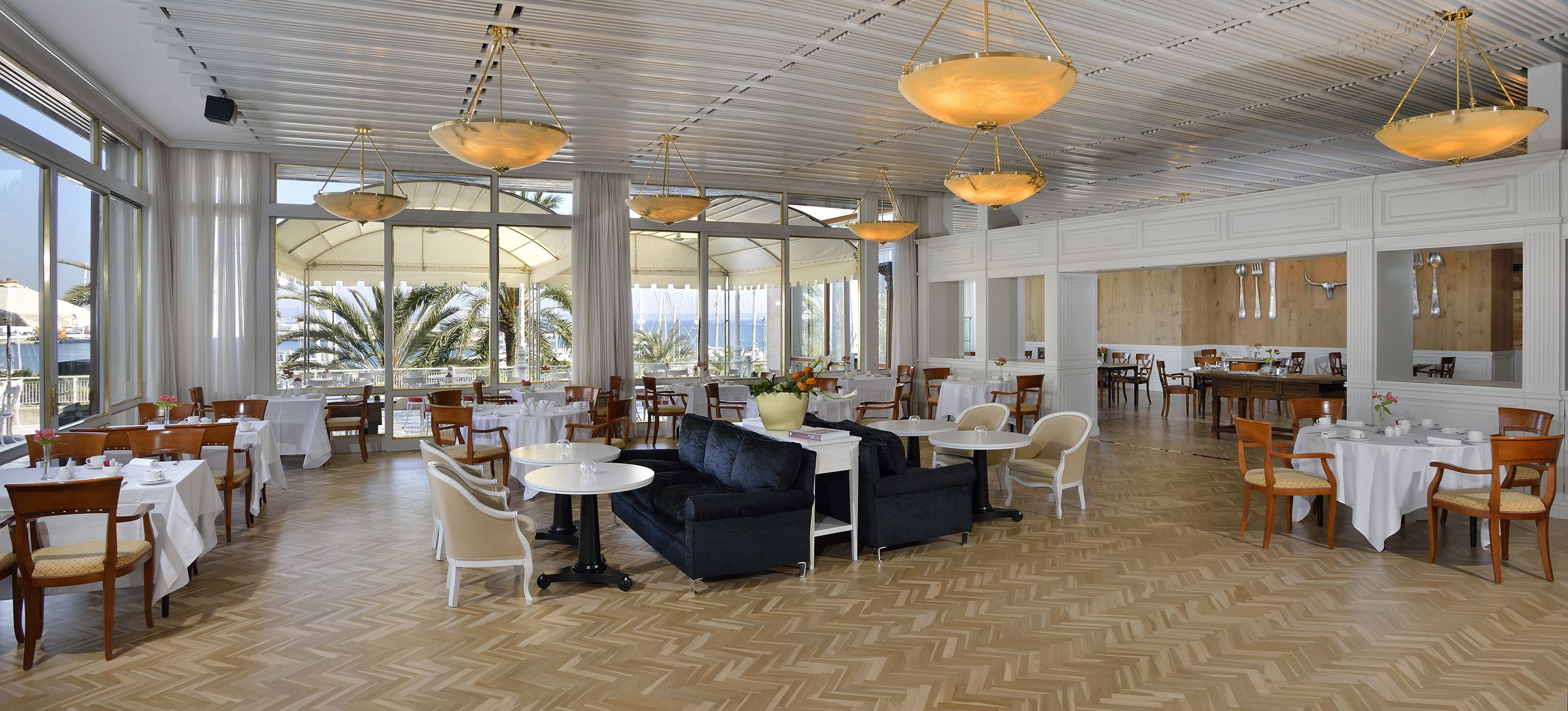 A spacious hotel lobby with multiple dining areas, large windows offering views of palm trees, and a modern chandelier.