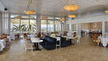 A spacious hotel lobby with multiple dining areas, large windows offering views of palm trees, and a modern chandelier.