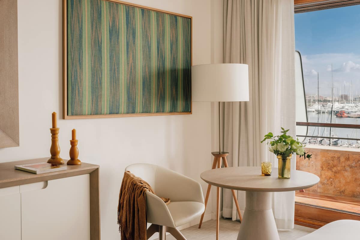 Un hotel moderno con un cuadro abstracto de gran tamaño, una mesa redonda con un florero, una silla con una manta y vistas a barcos a través de la ventana.