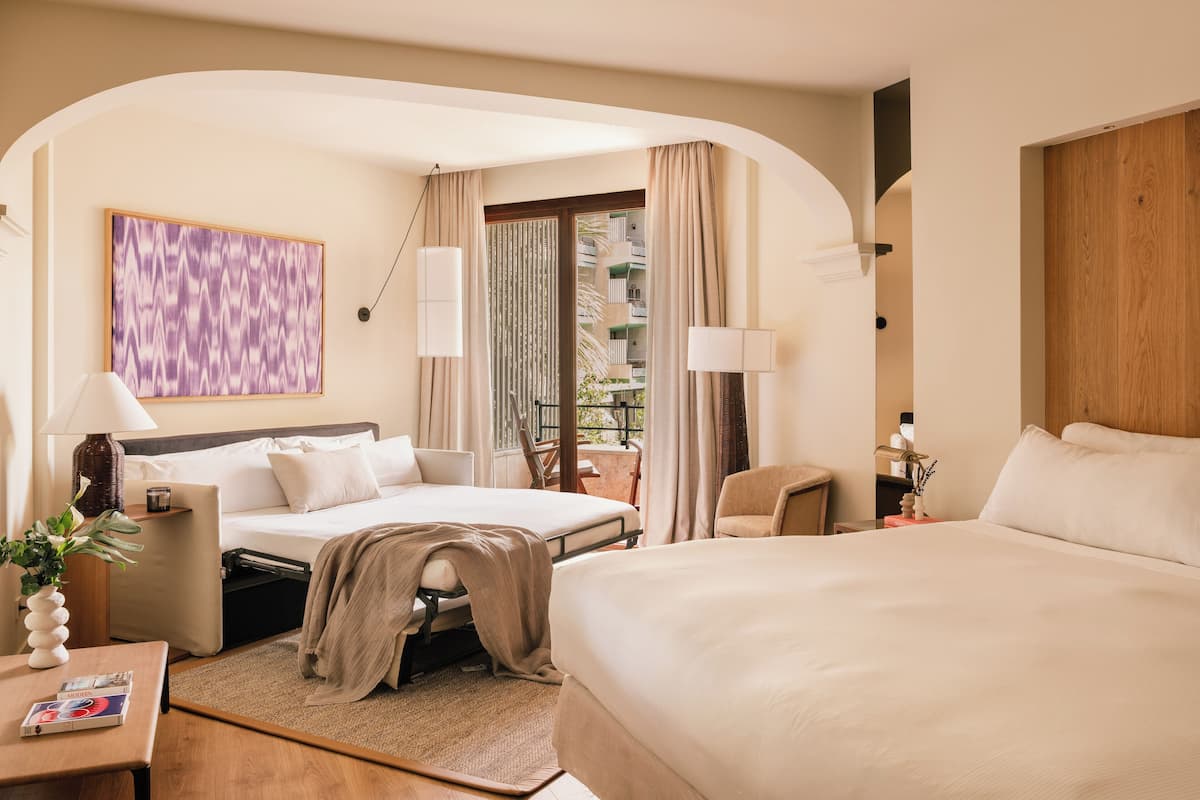 Habitación de hotel con dos camas, un escritorio, una silla, ventana con cortinas, vista al balcón y un cuadro abstracto moderno.