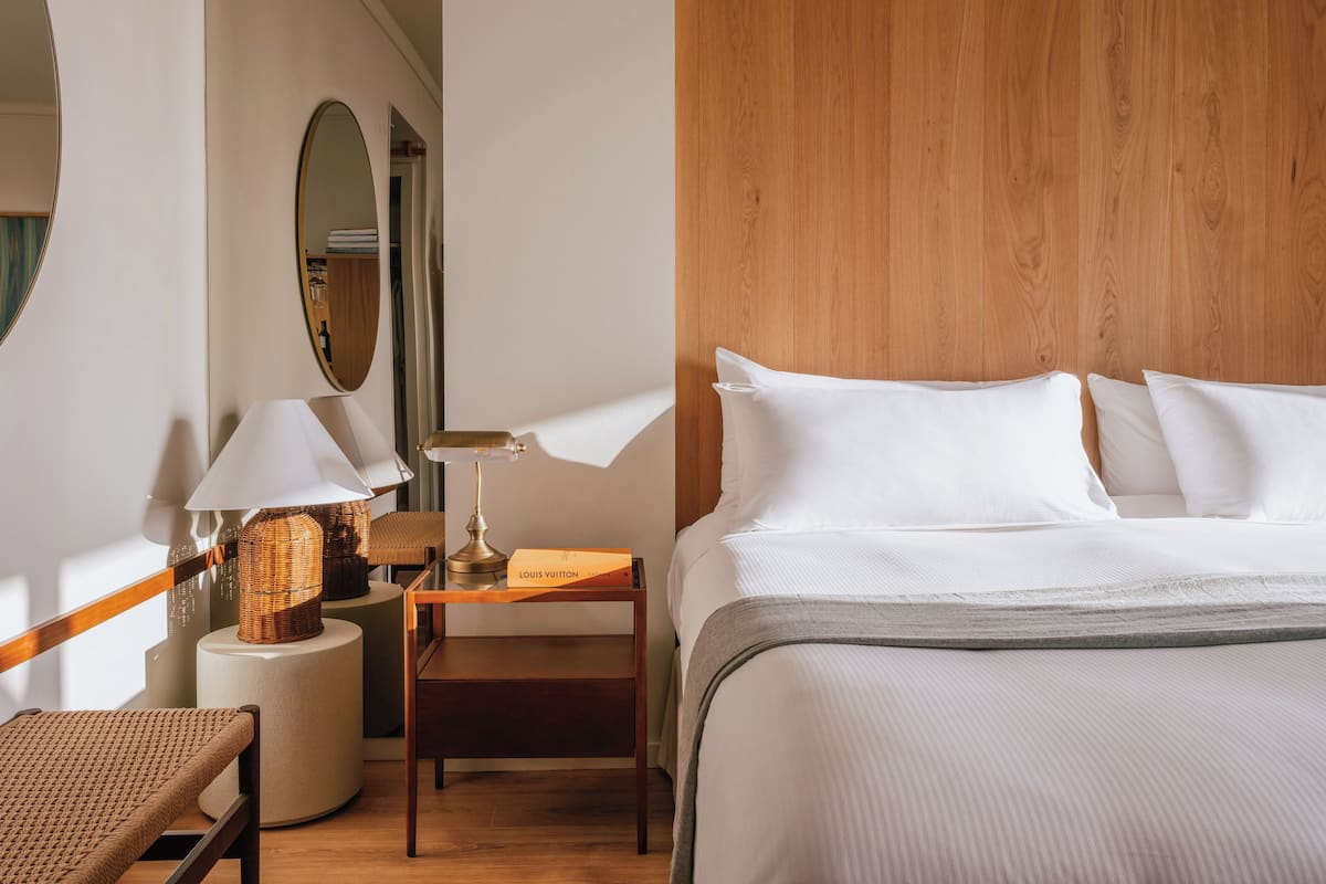 Habitación de hotel con cabecera de madera, cama con ropa blanca, mesita de noche con lámpara, silla de mimbre y una mesita auxiliar redonda con lámpara y una caja de Louis Vuitton.