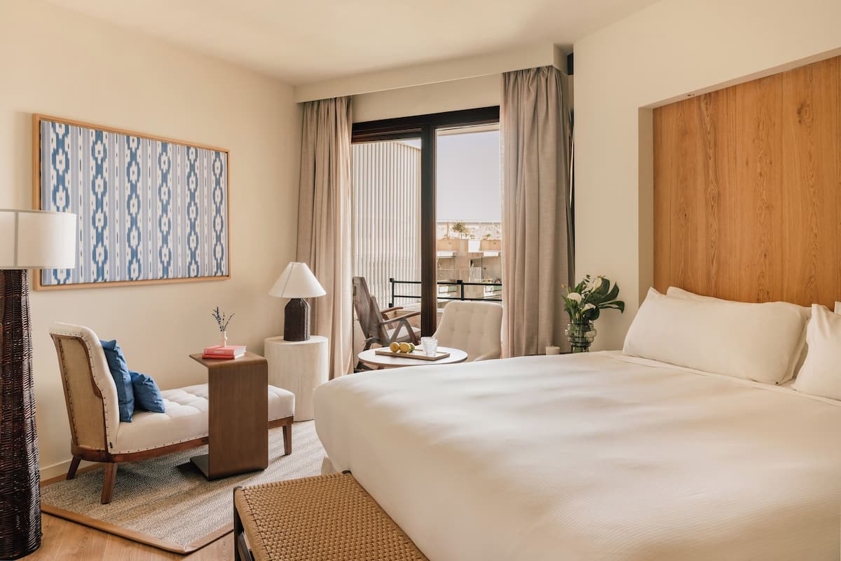 Habitación de hotel con una cama grande, una silla, una mesita con lámpara, un jarrón decorativo y vistas al exterior.
