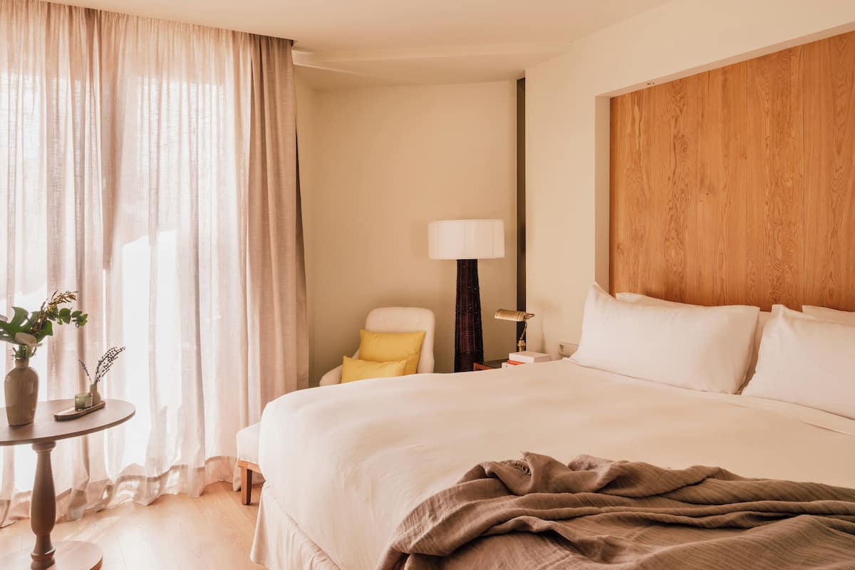 Una habitación de hotel con una cama grande, una mesita de noche con lámpara, una silla y una ventana con cortinas translúcidas.