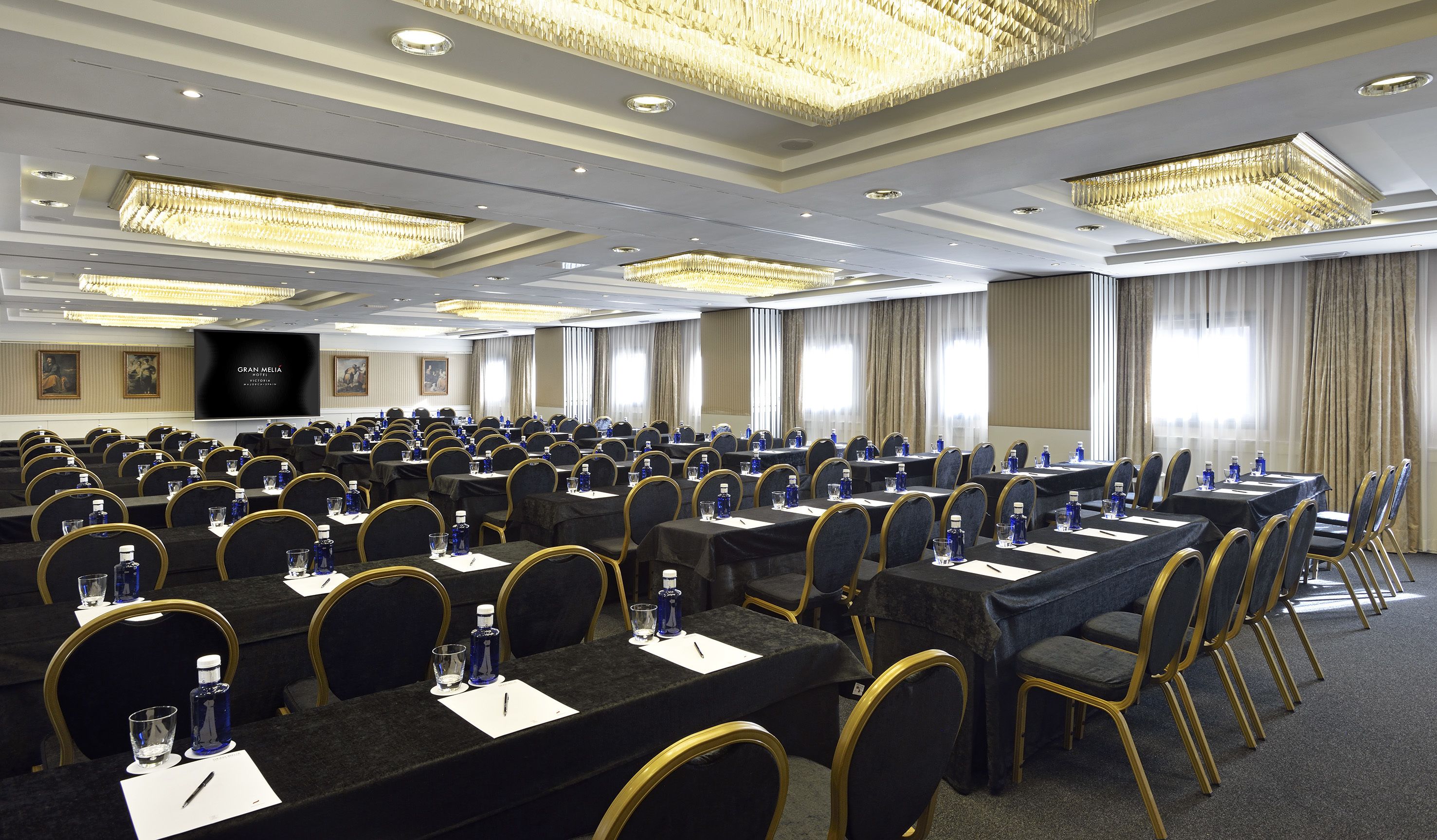A large conference room with rows of tables and chairs, equipped with water bottles and name cards, and a large screen at the front.