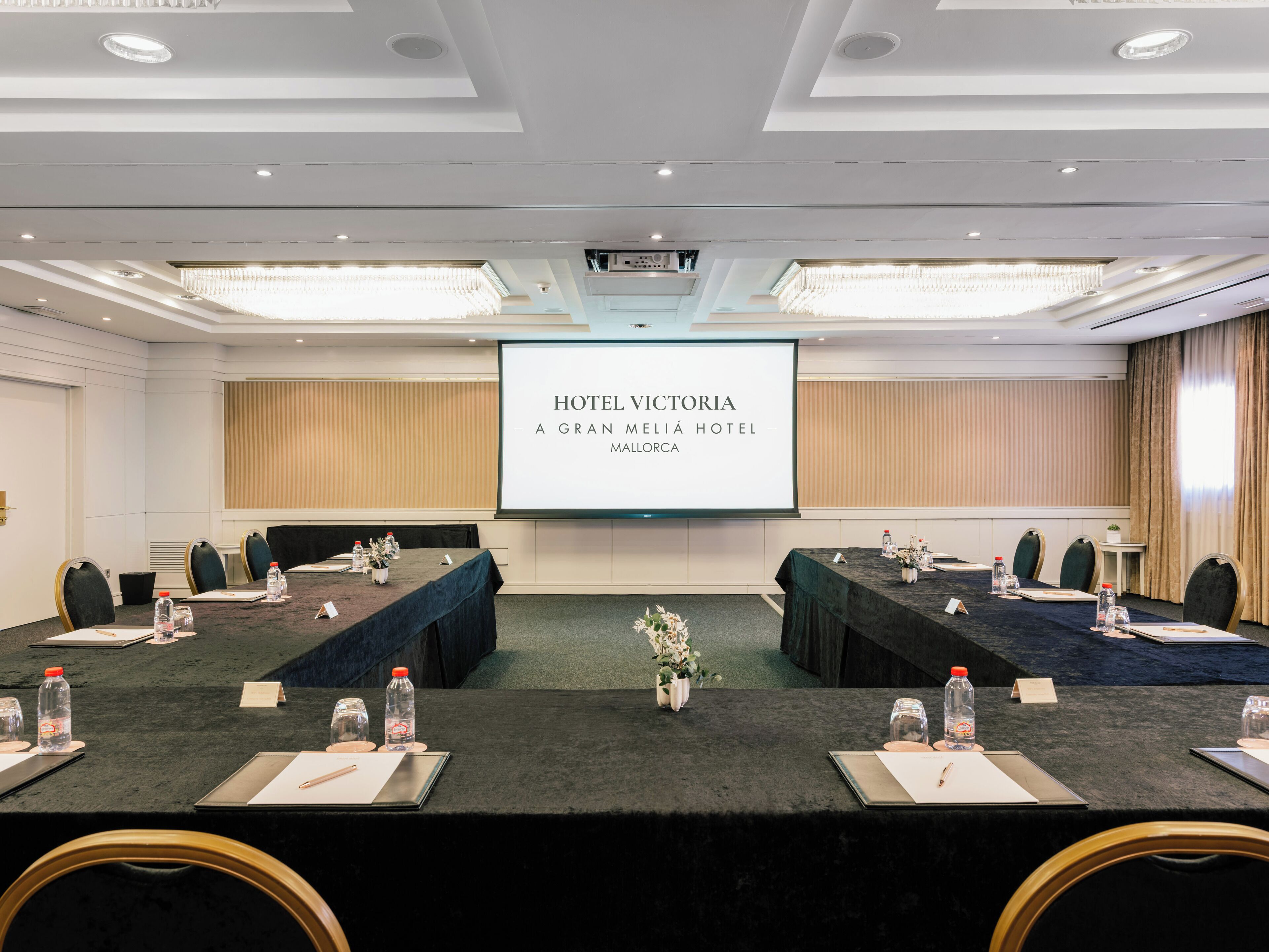 A conference room at Hotel Victoria with a large screen displaying the hotel's name, a long table with chairs, water bottles, and name cards.