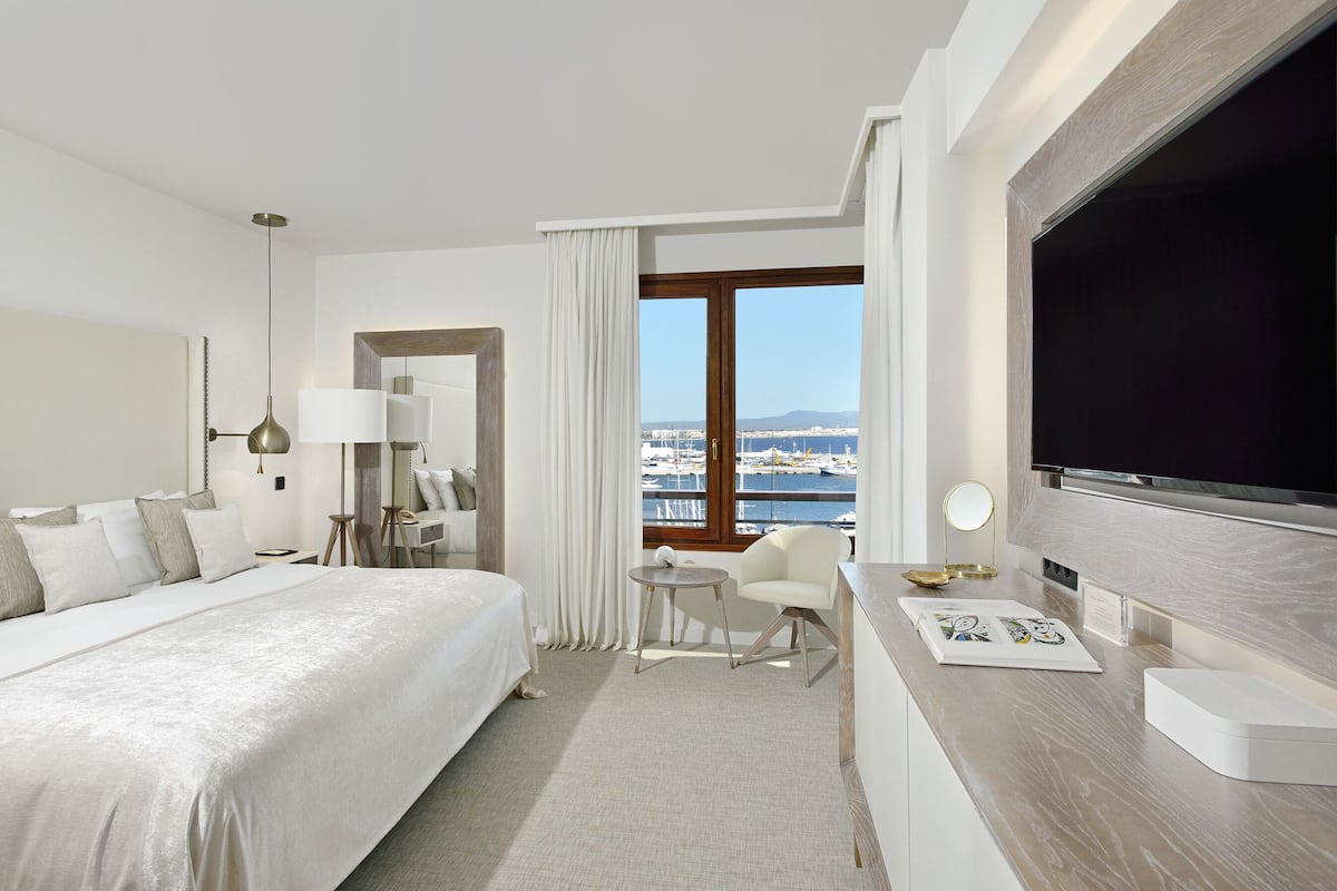 Habitación de hotel con una cama grande, un escritorio con televisión de pantalla plana, una mesita con lámpara y vista al puerto.