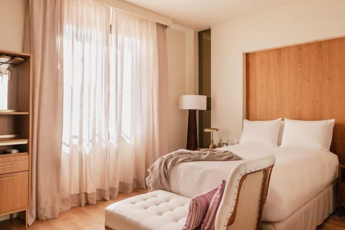 Une chambre d’hôtel avec un grand lit, une table de chevet, une lampe et une fenêtre avec des rideaux légers.