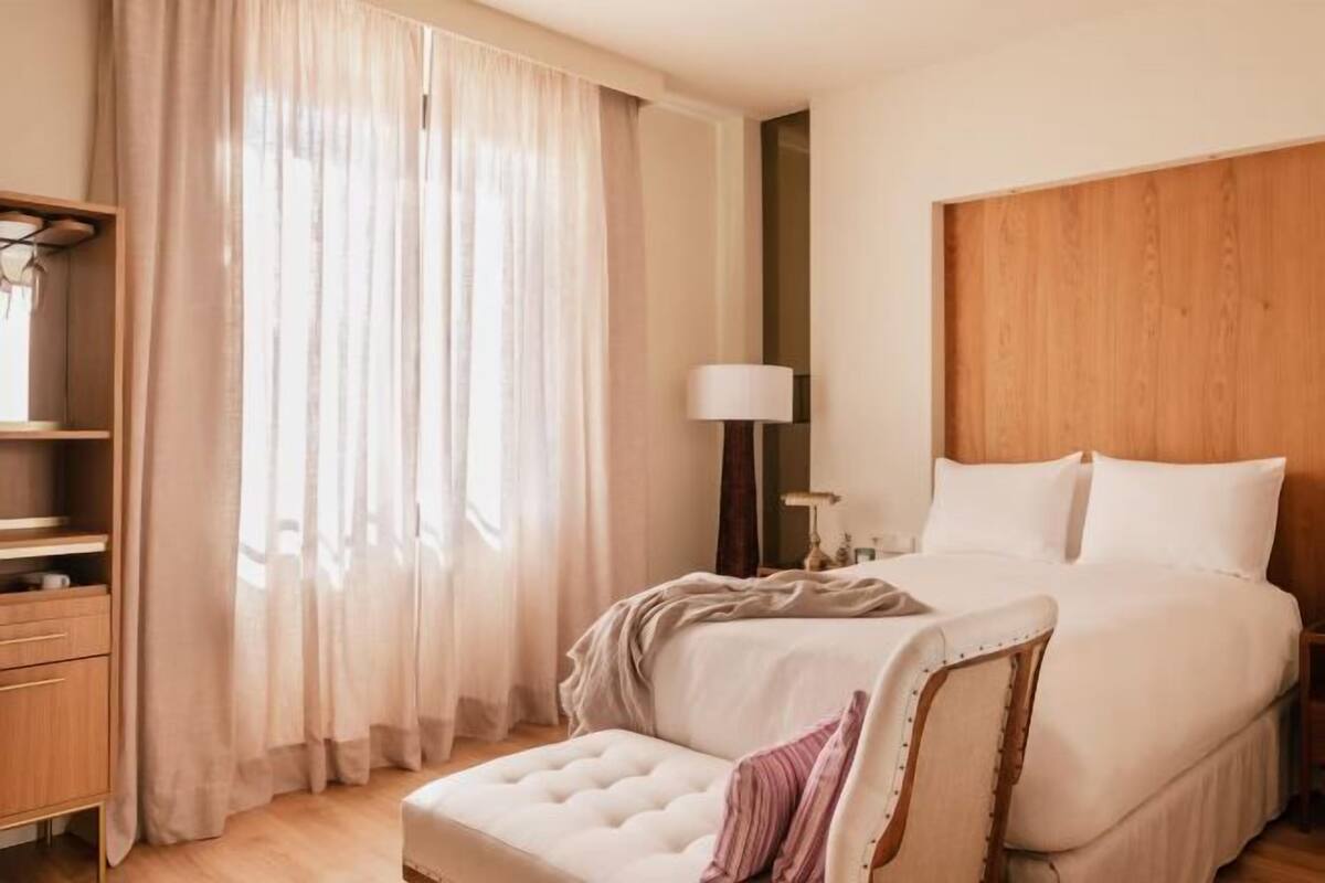 Un dormitorio con cama, mesita de noche, lámpara y ventana con cortinas.