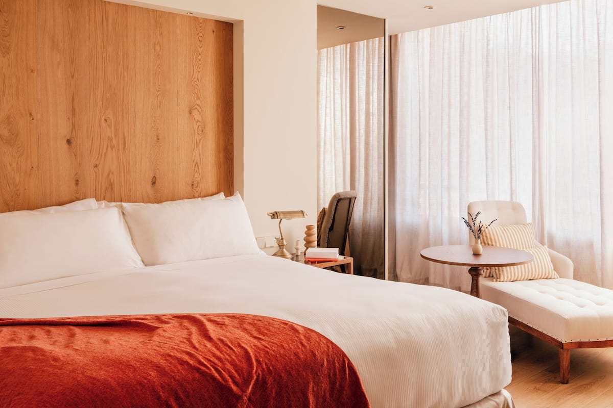 Habitación de hotel con una cama grande, cabecera de madera, una mesita redonda con una lámpara, una silla y vistas al exterior a través de cortinas traslúcidas.