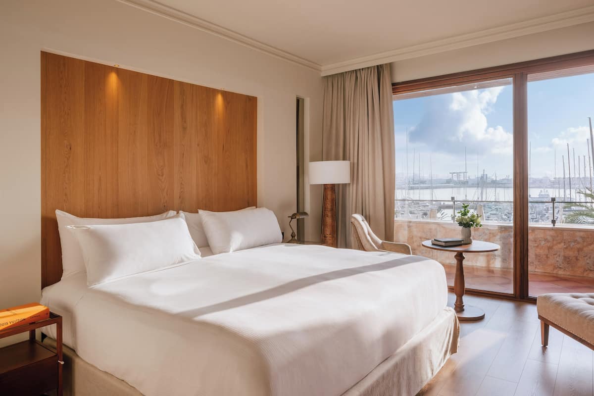 Habitación de hotel con una cama grande, una silla, una mesita con un florero y vistas al puerto deportivo.
