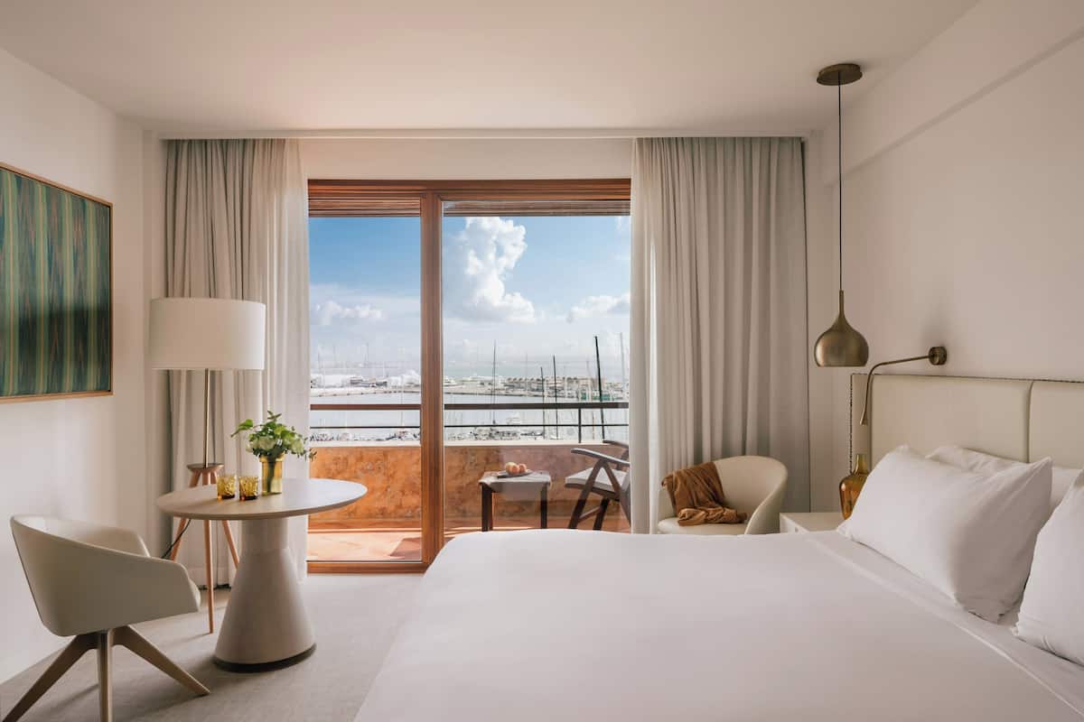 Una habitación de hotel con una cama grande, una mesa redonda con un jarrón de flores, una silla, una mesita y vistas al puerto deportivo a través de una puerta corrediza.