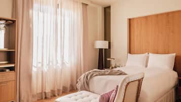 A hotel room with a bed, a bedside table with a lamp, a wooden headboard, a window with sheer curtains, and a wooden nightstand.