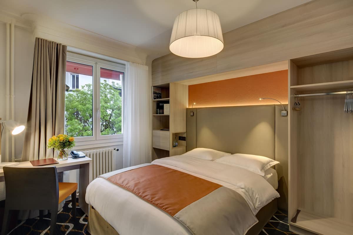 A hotel room with a bed, a desk, a chair, a lamp, and a window with curtains.