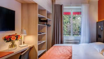 Quarto de hotel com cama, escrivaninha com luminária, estante e janela com cortinas.
